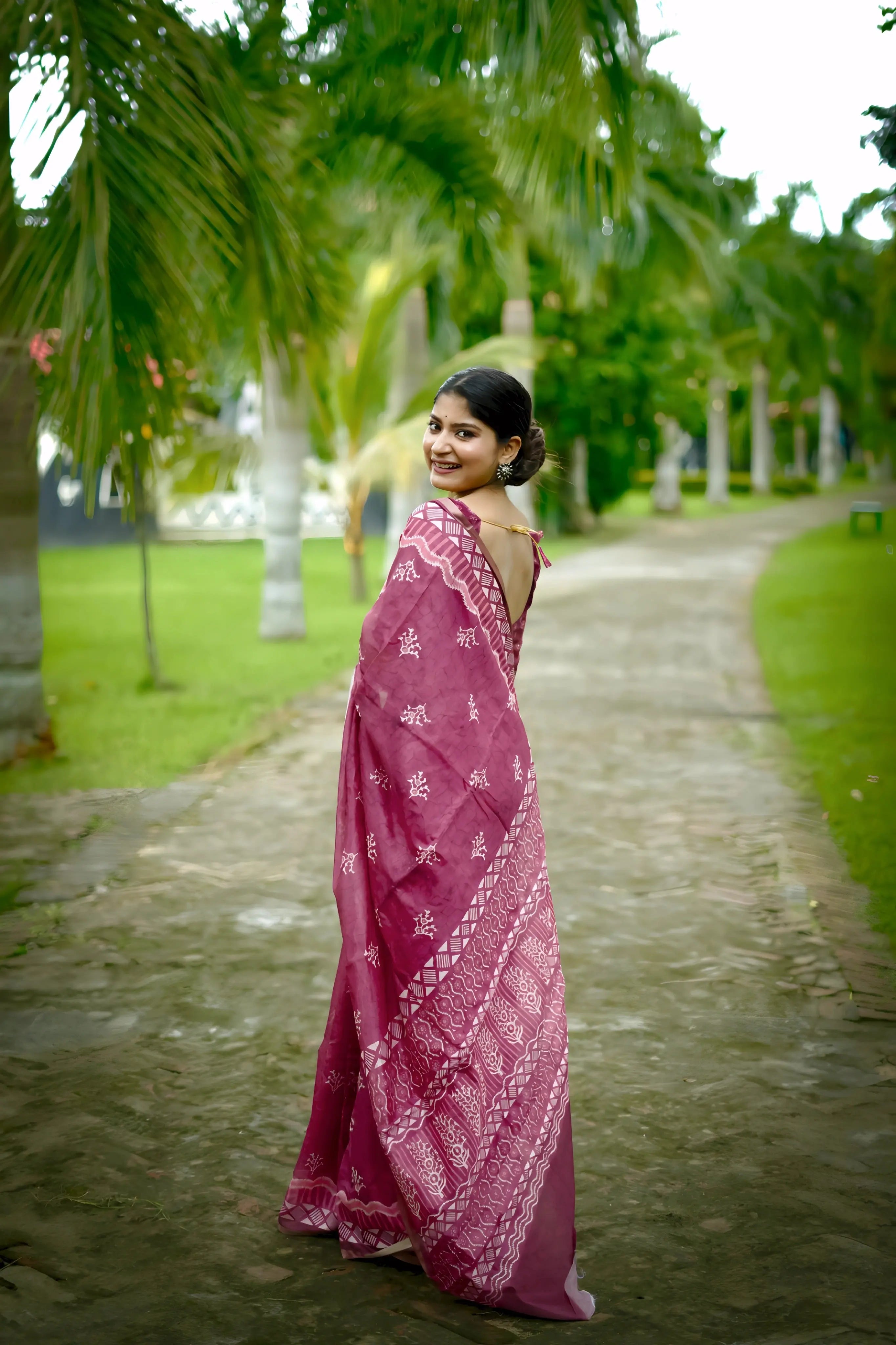 A woman models a Dusty Rose Pink Soft Cotton Saree featuring an all-over digital block print of traditional motifs. She wears a sheer-sleeved pink blouse and silver jhumka earrings, posing on an outdoor path lined with palm trees.